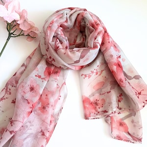 May include: A sheer scarf featuring a floral design in pink and red hues. The scarf is draped, displaying a watercolour-style pattern of cherry blossoms. A sprig of pink flowers is visible in the corner, adding to the aesthetic.