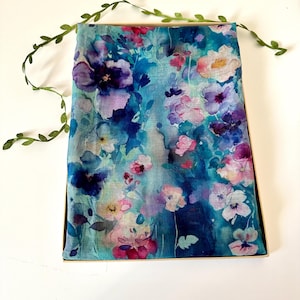 May include: A rectangular scarf featuring a watercolour-style floral print in shades of deep blue, turquoise, light purple, and soft pink. The fabric displays a vibrant, blended pattern of various flower shapes and foliage, neatly folded inside a box.