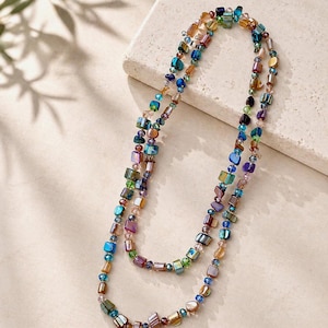 May include: Two long necklaces with colourful, iridescent beads. The beads are a mix of square and round shapes in shades of blue, green, purple, and gold. The necklaces are displayed on a neutral surface.
