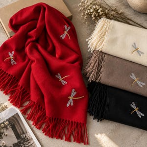 May include: A collection of soft scarves featuring embroidered dragonfly motifs. The set includes a vibrant red scarf, a cream scarf, a taupe scarf, and a black scarf. Each scarf is finished with long, delicate fringe along the bottom edges.