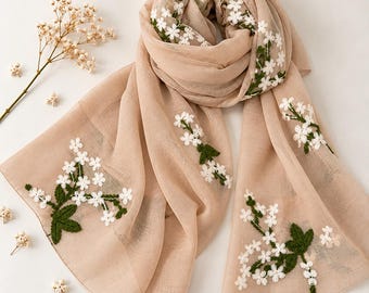 Op de afbeelding: Een beige sjaal met delicate witte bloemenborduursels, groene stengels en bladeren. De stof heeft een zachte, semi-transparante textuur. Het bloemmotief is verspreid over de sjaal met clusters van witte bloemen en groen bladwerk.