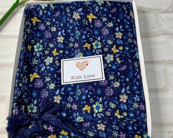 Op de afbeelding: Een marineblauwe sjaal met een bloemenprint en kwastjes, verpakt in een witte cadeaudoos met een hartvormig label waarop "With Love" staat.