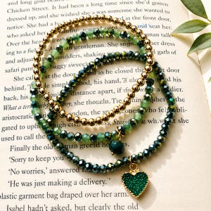 May include: Three layered bracelets. The top bracelet features gold beads. The middle and bottom bracelets have green beads. The bottom bracelet has a green heart charm. The bracelets are displayed on a book.