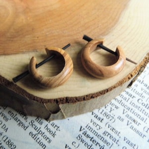 May include: Two wooden hoop earrings with a natural wood grain. Each earring features a curved, circular design with a black post. The earrings are displayed on a wooden surface, likely a slice of wood.