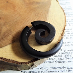 May include: A pair of black wooden spiral ear gauges. The gauges are made of dark wood and have a spiral design. They are shown on a wooden surface.
