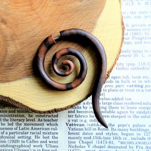 May include: A wooden spiral ear gauge, also known as a plug or tunnel, is made of dark brown wood with a reddish-brown swirl pattern. The gauge is resting on a wooden surface.