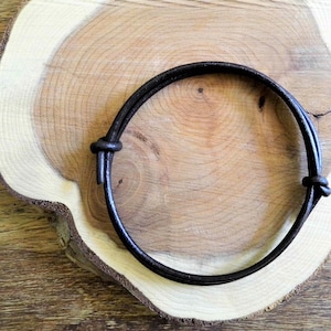 Może przedstawiać: Ciemnobrązowa skórzana bransoletka z wiązanym zapięciem. Bransoletka jest okrągła i wydaje się regulowana. Prezentowana jest na jasnobrązowej drewnianej powierzchni z naturalnym usłojeniem.