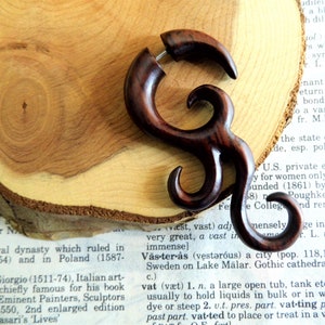 May include: A dark brown wooden ear gauge with a unique, intricate design. The gauge is shaped like a spiral with multiple curves and loops. It is a handmade piece of jewellery that is perfect for anyone who wants to add a touch of style to their look.