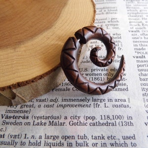May include: A wooden spiral ear gauge with a brown wood finish and a geometric pattern. The gauge is carved with a triangular design.
