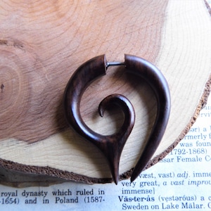 May include: A dark brown wooden ear gauge with a spiral design. The gauge is on a wooden surface.