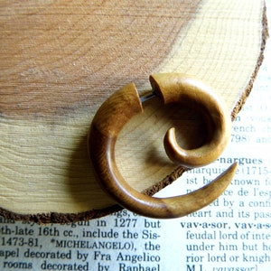 May include: A wooden spiral ear gauge, also known as a flesh tunnel, is shown on a wooden surface. The ear gauge is a light brown color and has a smooth, polished finish.