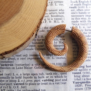 May include: A pair of wooden earrings with a carved wave pattern. The earrings are a light brown color and have a natural wood finish.
