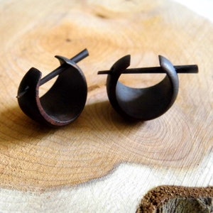 May include: A pair of black wooden hoop earrings with a metal post and a small metal bar that goes through the hoop. The earrings are on a wooden surface.