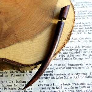 May include: A wooden ear gauge with a dark brown finish. The gauge is made of two pieces, a long, tapered piece and a shorter, cylindrical piece. The gauge is resting on a piece of wood with a natural grain pattern.