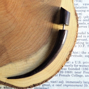 May include: A dark brown wooden ear gauge with a curved shape. The gauge is displayed on a wooden surface.