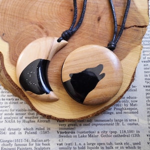 May include: Two wooden pendant necklaces. One is crescent-shaped with a black inlay and white speckles. The other is round with a howling wolf silhouette. Both have black cords. The pendants are on a wooden surface.