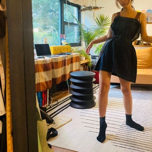 Può includere: Una donna che indossa un abito nero senza maniche con una cintura e calze nere. È in piedi in una stanza con una tovaglia a quadri, uno sgabello e una finestra sullo sfondo.