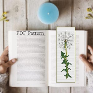 May include: A handmade bookmark featuring a cross-stitch design of a dandelion. The bookmark is green and white, with a detailed image of the dandelion's seed head and stem. The text "PDF Pattern" is visible on the open book.