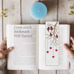 Puede incluir: Un patrón PDF de punto de cruz para marcapáginas con un diseño de pájaros en una rama con corazones rojos colgantes. El marcapáginas está en un libro abierto. El texto "Cross stitch bookmark PDF Pattern" es visible.