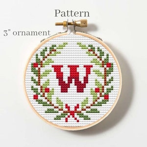 Puede incluir: Un aro de bordado redondo de 7,6 cm con un patr&oacute;n de punto de cruz de una letra "W" roja rodeada de una corona verde con bayas rojas. El patr&oacute;n se titula "Pattern".