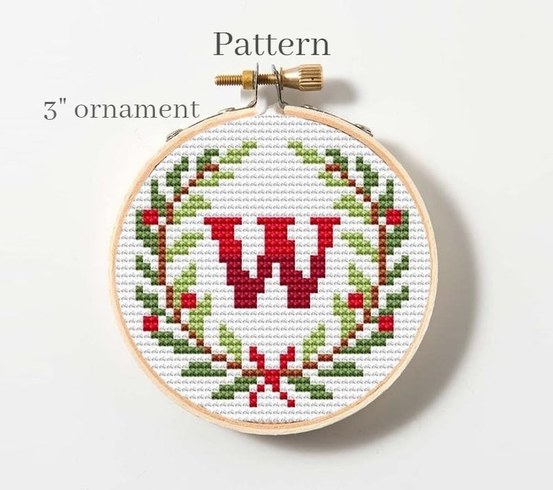 Puede incluir: Un aro de bordado redondo de 7,6 cm con un patr&oacute;n de punto de cruz de una letra "W" roja rodeada de una corona verde con bayas rojas. El patr&oacute;n se titula "Pattern".