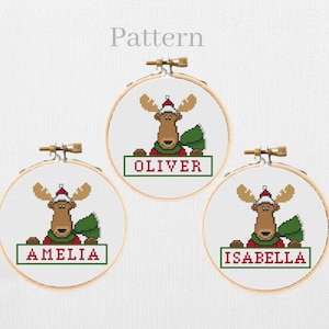 May include: Three circular wooden embroidery hoops containing cross-stitch patterns of a brown moose wearing a red and white Santa hat and a green scarf. Each hoop features a rectangular box with the names Oliver, Amelia, or Isabella in red.