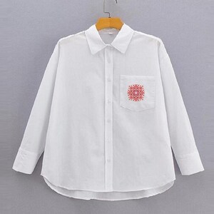 Puede incluir: Una camisa blanca abotonada con mangas largas y un bolsillo en el pecho. El bolsillo presenta un dise&ntilde;o de copo de nieve rojo. La camisa est&aacute; colgada en una percha de madera sobre un fondo neutro.