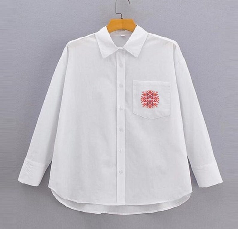 Puede incluir: Una camisa blanca abotonada con mangas largas y un bolsillo en el pecho. El bolsillo presenta un dise&ntilde;o de copo de nieve rojo. La camisa est&aacute; colgada en una percha de madera sobre un fondo neutro.