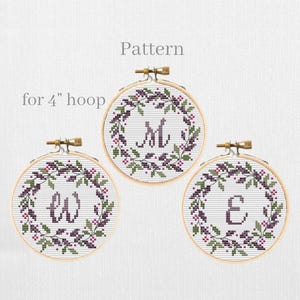May include: Three cross-stitch embroidery hoops, each with a letter (M, W, E) in a floral wreath design. The text "Pattern" and "for 4" hoop" are visible. The hoops are held in wooden frames.