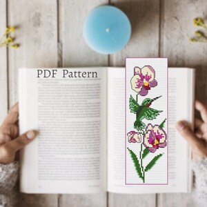 以下が含まれることがあります： ピンクと白の花のついたハチドリを特徴とするクロスステッチ刺繍のPDFパターン。パターンは本に表示されています。