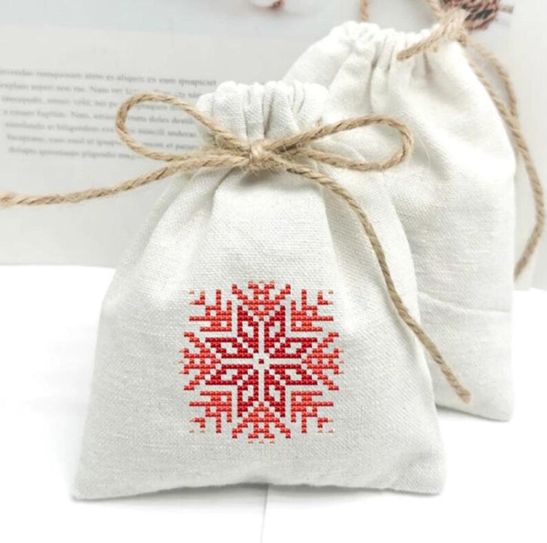 Puede incluir: Dos peque&ntilde;as bolsas con cord&oacute;n de color blanco roto. La bolsa delantera presenta un dise&ntilde;o de copo de nieve de punto de cruz rojo y blanco. Cada bolsa est&aacute; atada con un lazo de hilo natural. Las bolsas son de tela texturizada.