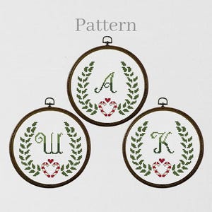 May include: Three round cross-stitch patterns with the letters A, W, and K. Each design features a green leaf wreath, a red and white heart, and a dark brown wooden hoop. The word "Pattern" is at the top.