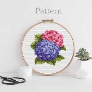Puede incluir: Un patrón de punto de cruz de dos hortensias, una rosa y otra morada, con hojas verdes. El patrón se muestra en un aro de bordado de madera.