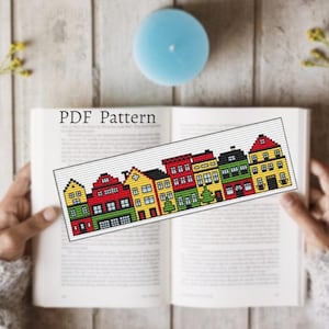 Könnte beinhalten: Ein Kreuzstich-Lesezeichen mit einer Reihe farbenfroher Häuser mit roten, gelben, grünen und schwarzen Dächern. Das Lesezeichen befindet sich in einem offenen Buch mit dem Text "PDF Pattern". Eine blaue Kerze und ein Zweig gelber Blumen sind im Hintergrund.