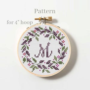 May include: A cross-stitch pattern in a 4-inch embroidery hoop. The design features a wreath of purple and green leaves and flowers surrounding the letters "M" and "L". The text "Pattern" and "for 4" hoop" are above and to the left of the hoop.