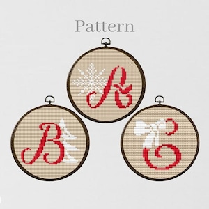 Puede incluir: Tres aros de bordado redondos con tela beige y bordado rojo y blanco. Los aros presentan las letras A, B y C en una fuente cursiva con un copo de nieve, un árbol de Navidad y un lazo.