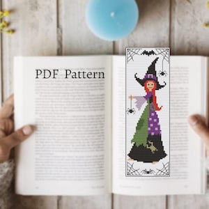 May include: A cross stitch pattern featuring a witch with red hair and a green and purple dress. The witch is holding a spider on a string. The pattern is surrounded by a black border with spiders and webs.