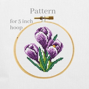 May include: A cross-stitch pattern of purple crocus flowers with green leaves centered in a round wooden embroidery hoop. The text Pattern for 5 inch hoop is printed in gray above the hoop, which features a metal tightening screw at the top.