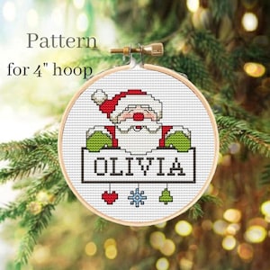 May include: A cross-stitch pattern for a 4-inch hoop featuring a Santa Claus holding a sign with the name "Olivia" on it. The sign is decorated with a heart, snowflake, and Christmas tree.