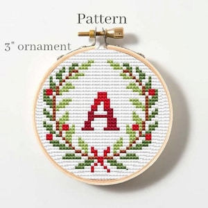 以下が含まれることがあります： 白い背景に赤い文字「A」が描かれた、緑色のリースと赤い実で飾られた直径7.6cmの丸いオーナメントです。