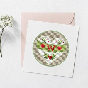 K&ouml;nnte beinhalten: Eine wei&szlig;e Karte mit einem gr&uuml;nen Kreis, der ein wei&szlig;es Herz mit einem gr&uuml;n-roten Design und dem Buchstaben "W" in der Mitte enth&auml;lt. Das Herz hat rote Herzen und gr&uuml;ne Stechpalmenbl&auml;tter.