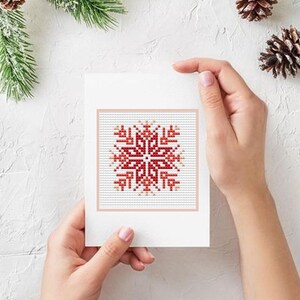 Puede incluir: Una tarjeta de felicitaci&oacute;n blanca con un dise&ntilde;o de copo de nieve rojo y blanco en punto de cruz. La tarjeta est&aacute; sostenida por manos, con pi&ntilde;as y vegetaci&oacute;n en el fondo, lo que sugiere un tema invernal o festivo.