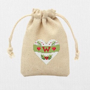 K&ouml;nnte beinhalten: Eine kleine, beige Kordeltasche mit einem wei&szlig;en Herz, das auf der Vorderseite gestickt ist. Das Herz hat einen gr&uuml;nen Rand und einen roten Buchstaben "W" in der Mitte. Um das Herz herum befinden sich kleine rote Herzen und gr&uuml;ne Stechpalmenbl&auml;tter.