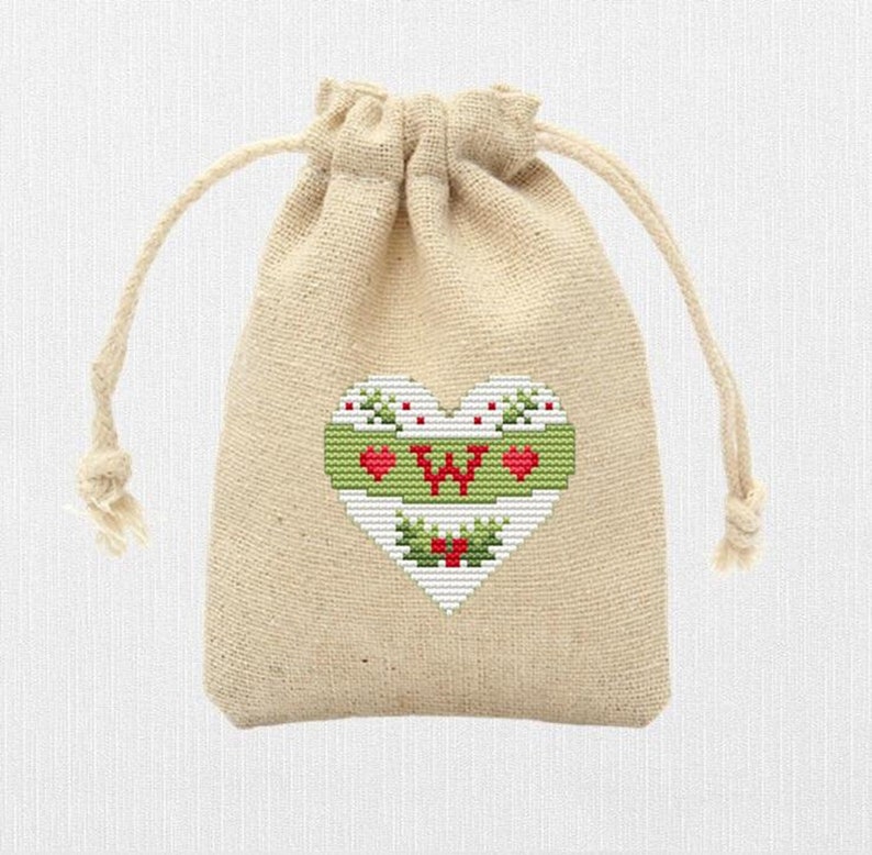 K&ouml;nnte beinhalten: Eine kleine, beige Kordeltasche mit einem wei&szlig;en Herz, das auf der Vorderseite gestickt ist. Das Herz hat einen gr&uuml;nen Rand und einen roten Buchstaben "W" in der Mitte. Um das Herz herum befinden sich kleine rote Herzen und gr&uuml;ne Stechpalmenbl&auml;tter.