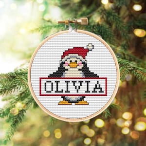 Può includere: Un ornamento bianco a punto croce con un pinguino che indossa un cappello rosso di Babbo Natale e tiene un cartello con su scritto "OLIVIA".