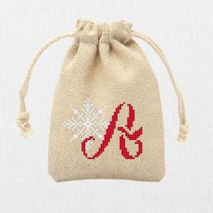 May include: A small, beige fabric drawstring bag with a red embroidered letter "A" and a white snowflake design.