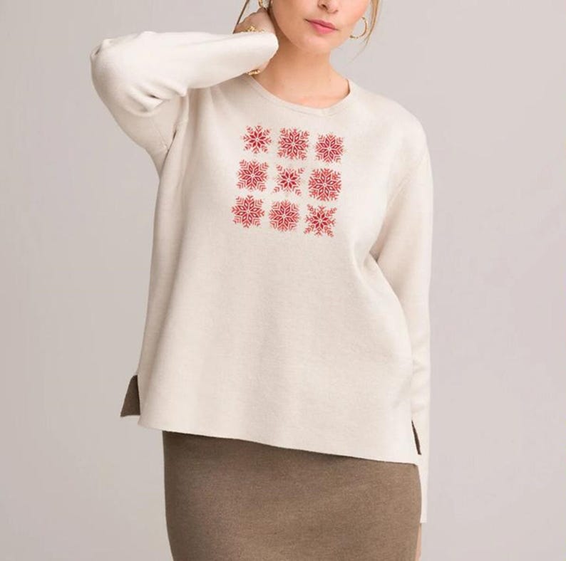 Puede incluir: Un su&eacute;ter de manga larga color crema con un patr&oacute;n cuadrado de nueve dise&ntilde;os de copos de nieve rojos en la parte delantera. El su&eacute;ter tiene un cuello redondo y aberturas laterales. La modelo lleva una falda marr&oacute;n.