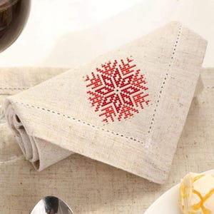 Puede incluir: Una servilleta de lino doblada con un dise&ntilde;o de copo de nieve bordado en rojo y blanco. La servilleta tiene un borde cosido decorativo y est&aacute; colocada sobre un mantel de color neutro. Un vaso de l&iacute;quido rojo es visible en el fondo.