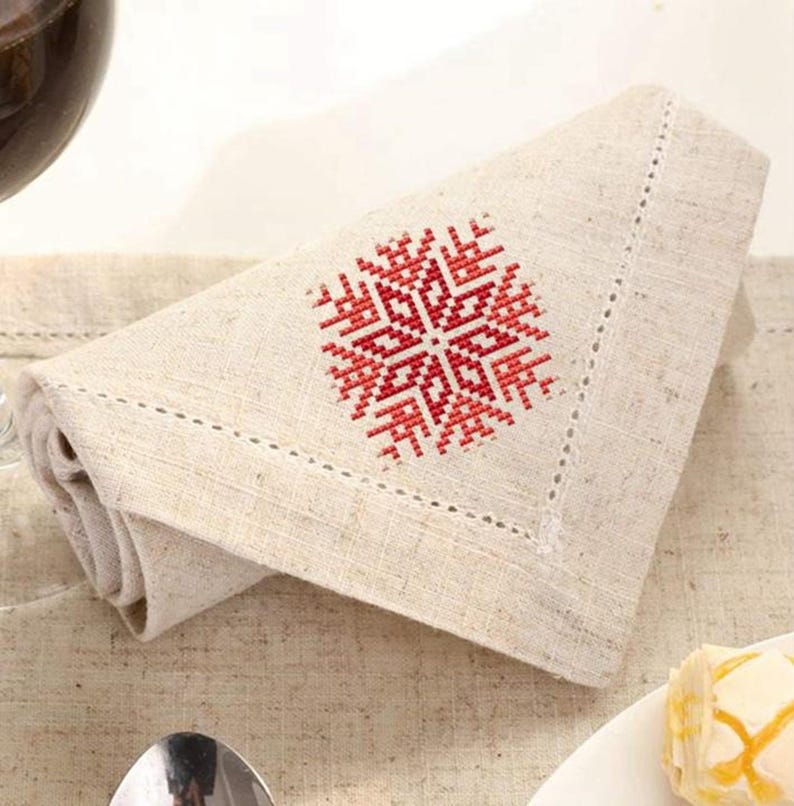 Puede incluir: Una servilleta de lino doblada con un dise&ntilde;o de copo de nieve bordado en rojo y blanco. La servilleta tiene un borde cosido decorativo y est&aacute; colocada sobre un mantel de color neutro. Un vaso de l&iacute;quido rojo es visible en el fondo.