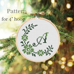 Puede incluir: Un patrón de punto de cruz en un aro de 10 cm. El diseño presenta una corona floral verde y la letra "A" en verde. El texto "Pattern for 4" hoop" está encima del diseño.